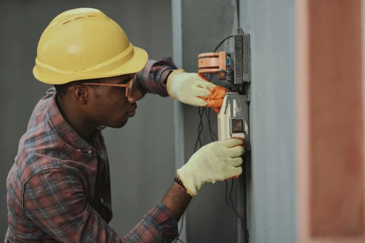 African American electrician in a yellow hardhat working on a wall electrical panel
