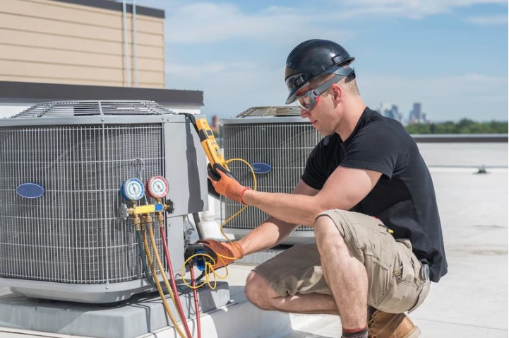 HVAC Technician in a black hardhat knelling and connecting gauges to a HVAC unit on a roof