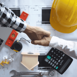 Two hands shaking above a table that has draft plans, level, calculator, and yellow hard hat.