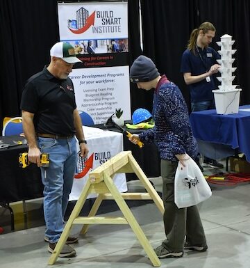 Build Smart Instructor stands in the Build Smart booth at expo as a student hammers a nail into a board on a sawhorse.