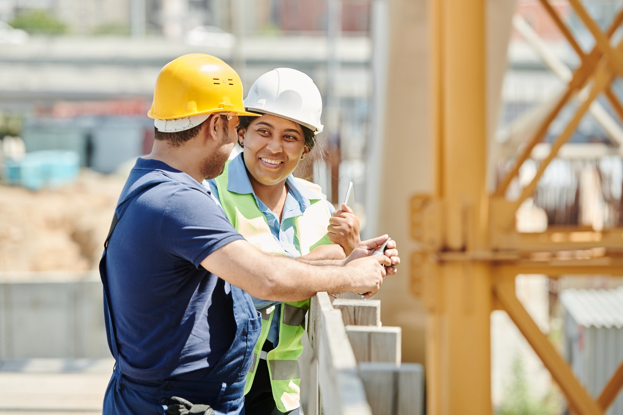 Two construction workers in hard hats smiling and talking on a construction site